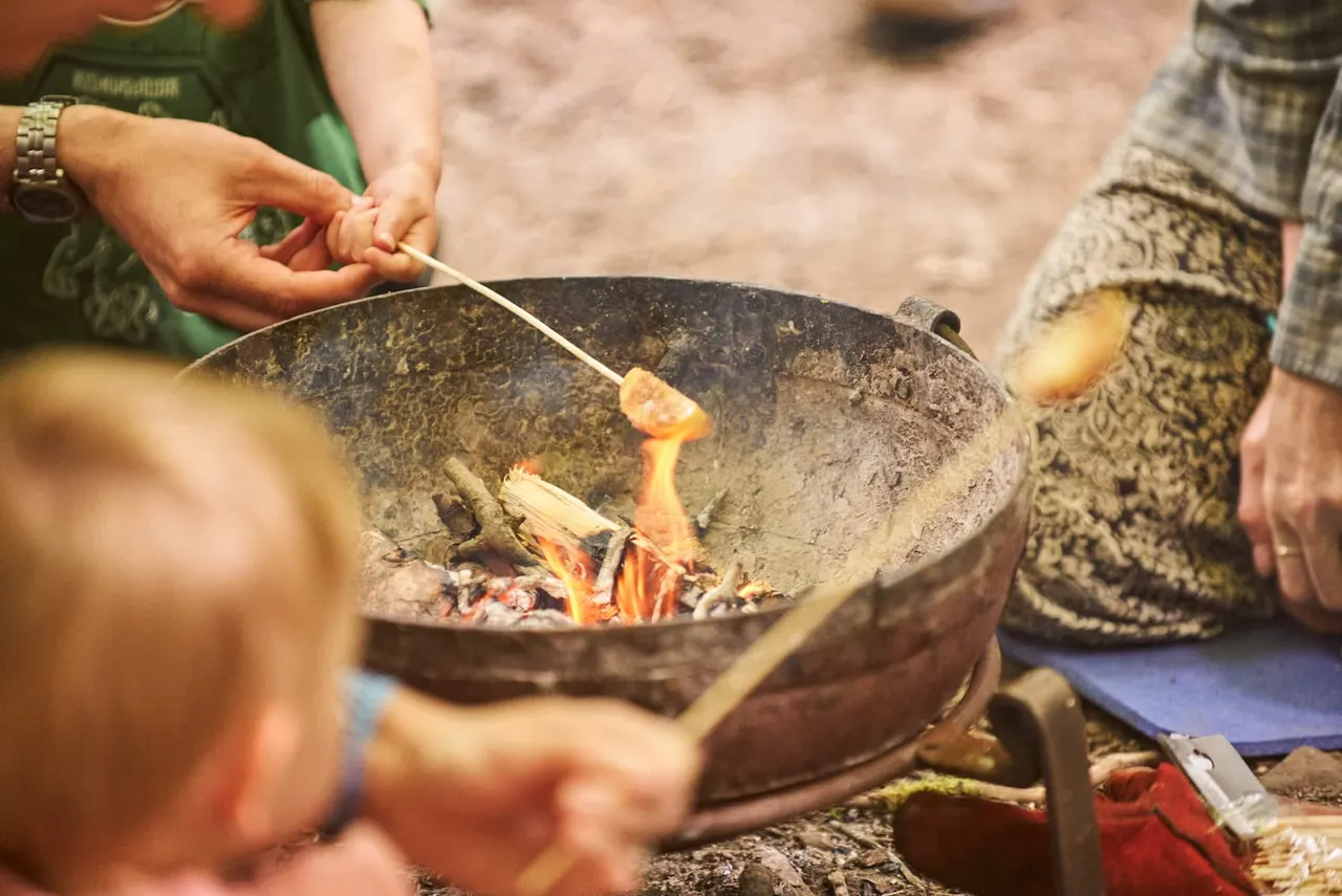 Person cooking food on a stick over an open fire in a large metal bowl outdoors.