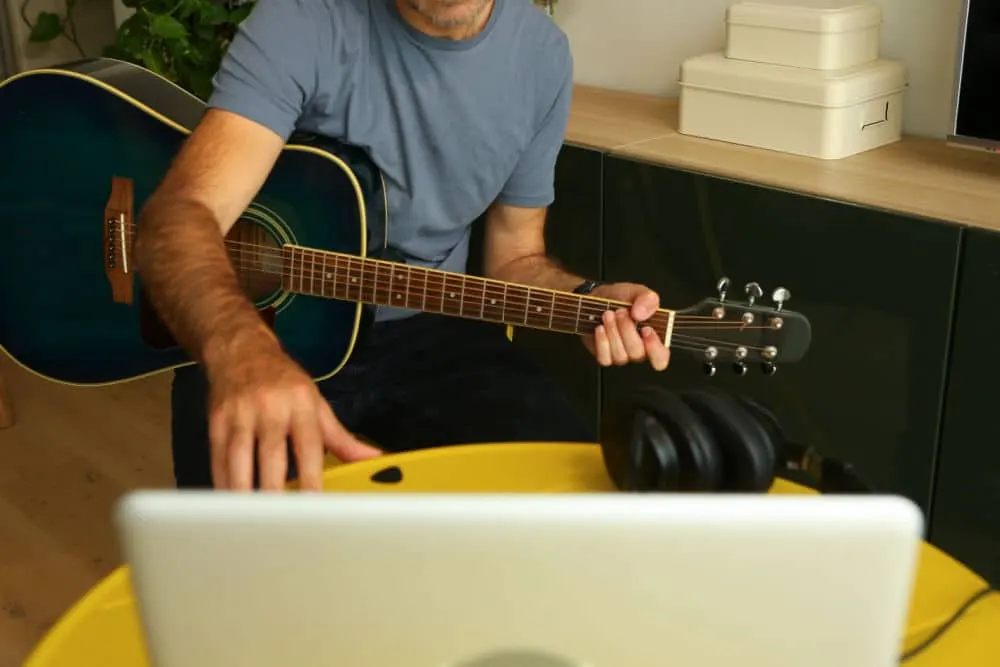 Man in blue t-shirt playing acoustic guitar whilst seated at desk with laptop.