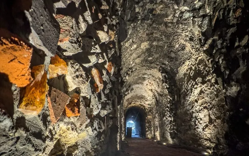 Ancient stone tunnel with weathered walls lit by warm and cool lighting, leading towards a doorway with blue light in the distance.