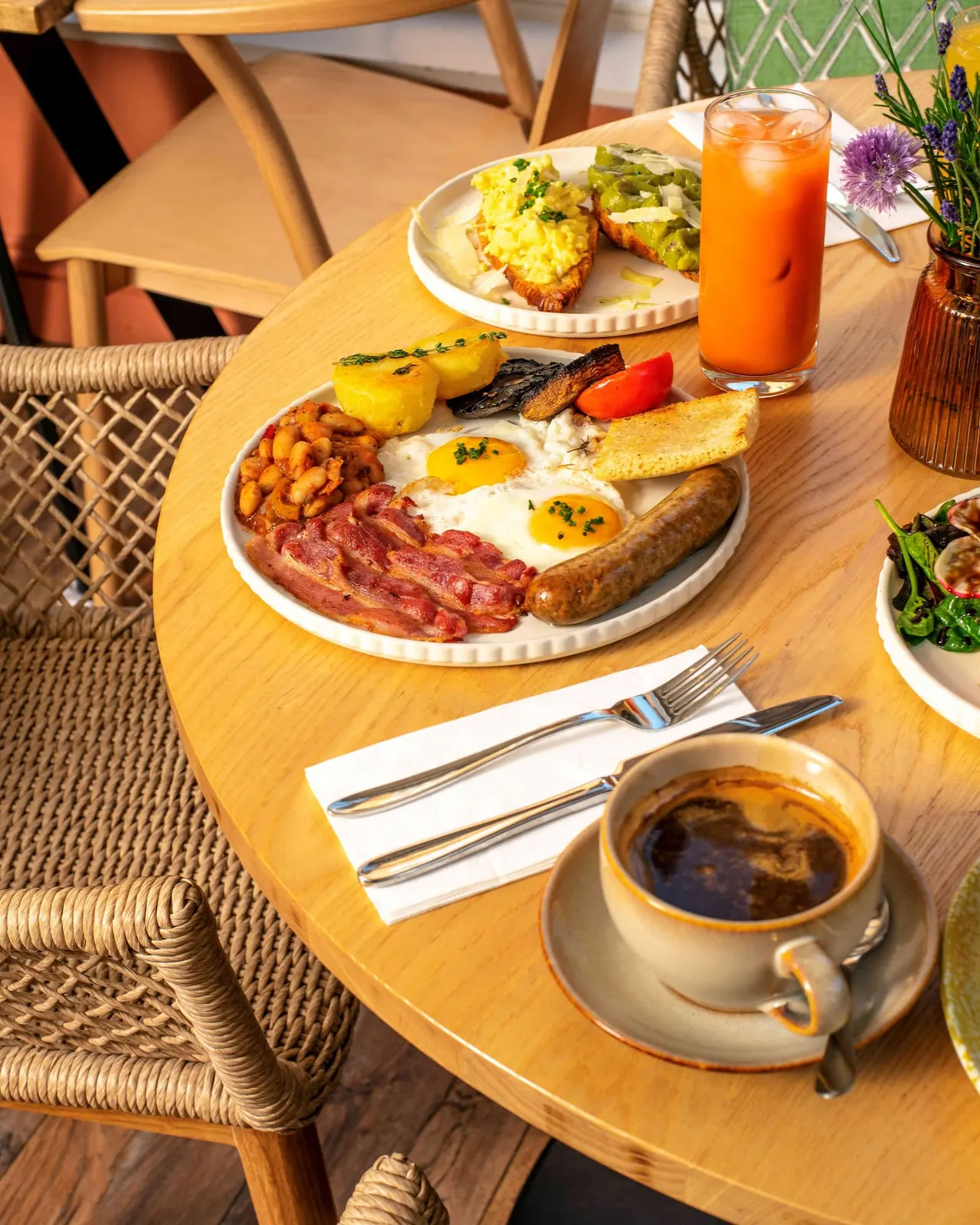 Full breakfast spread on a wooden table with eggs, bacon, sausage, toast, and side dishes, with coffee and orange juice.