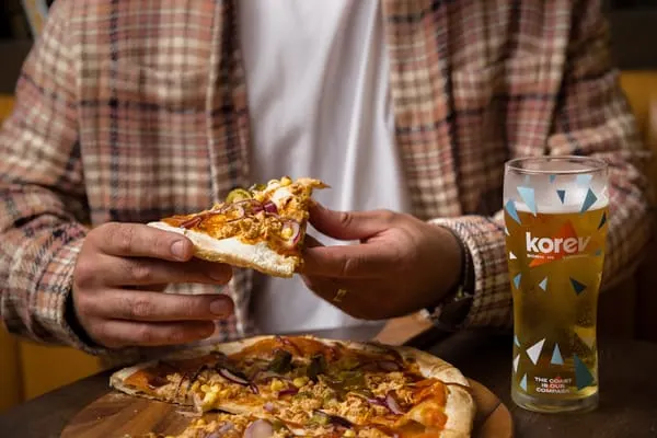 Person in checked shirt holding a slice of pizza with a branded drink on the table.