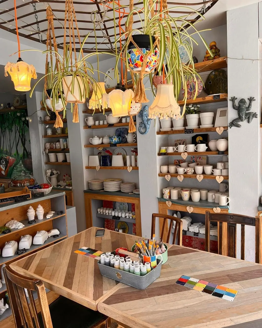 Pottery studio interior with wooden work table, art supplies, hanging plants, and shelves displaying ceramic pots and decorative items.