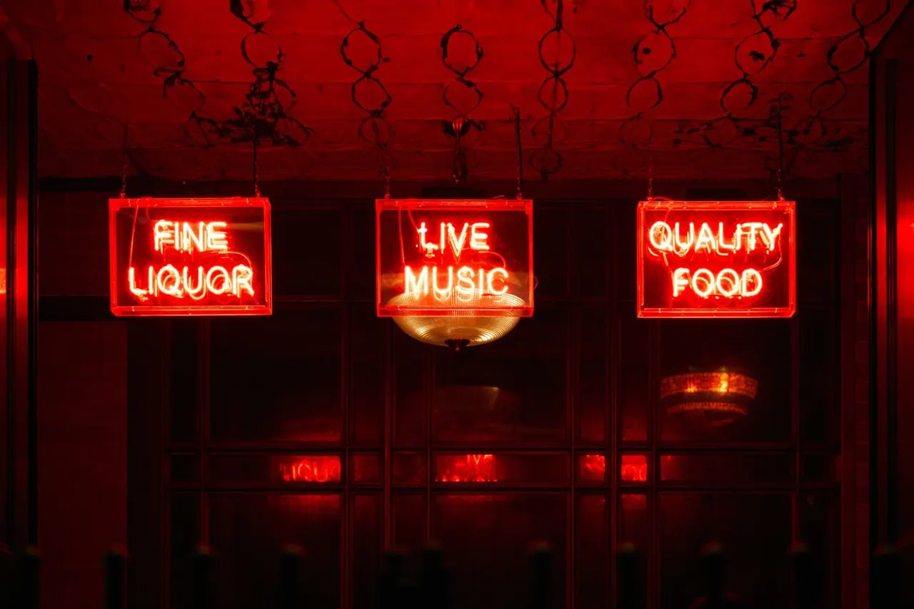 Red neon signs advertising fine liquor, live music, and quality food hang above a bar window at night.