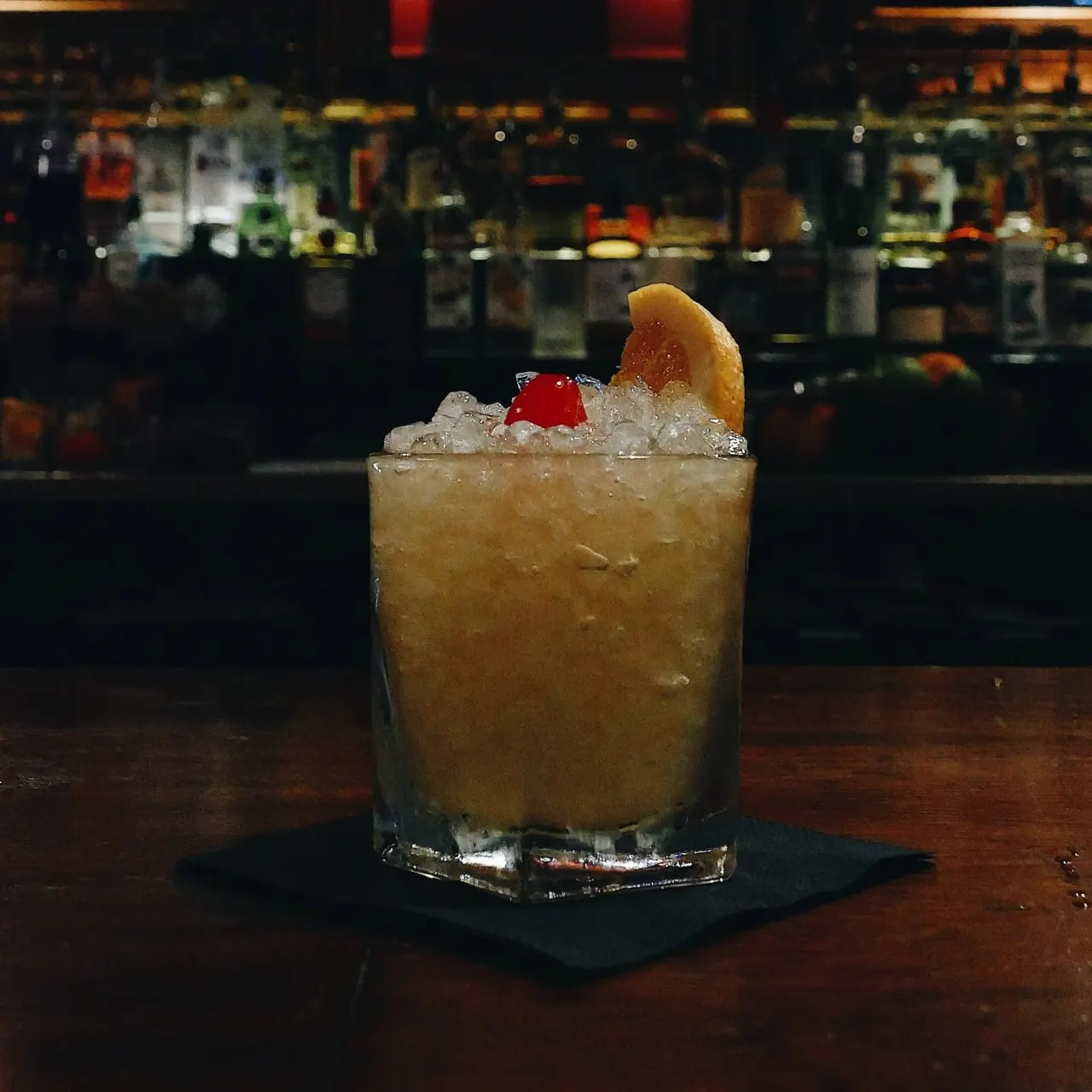 Cocktail with ice, orange slice and cherry garnish on a bar counter with blurred bar interior behind.