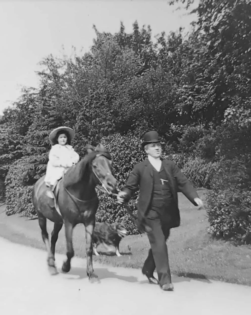 Woman in white dress and hat on horseback with man in dark suit and bowler hat leading horse along tree-lined path.