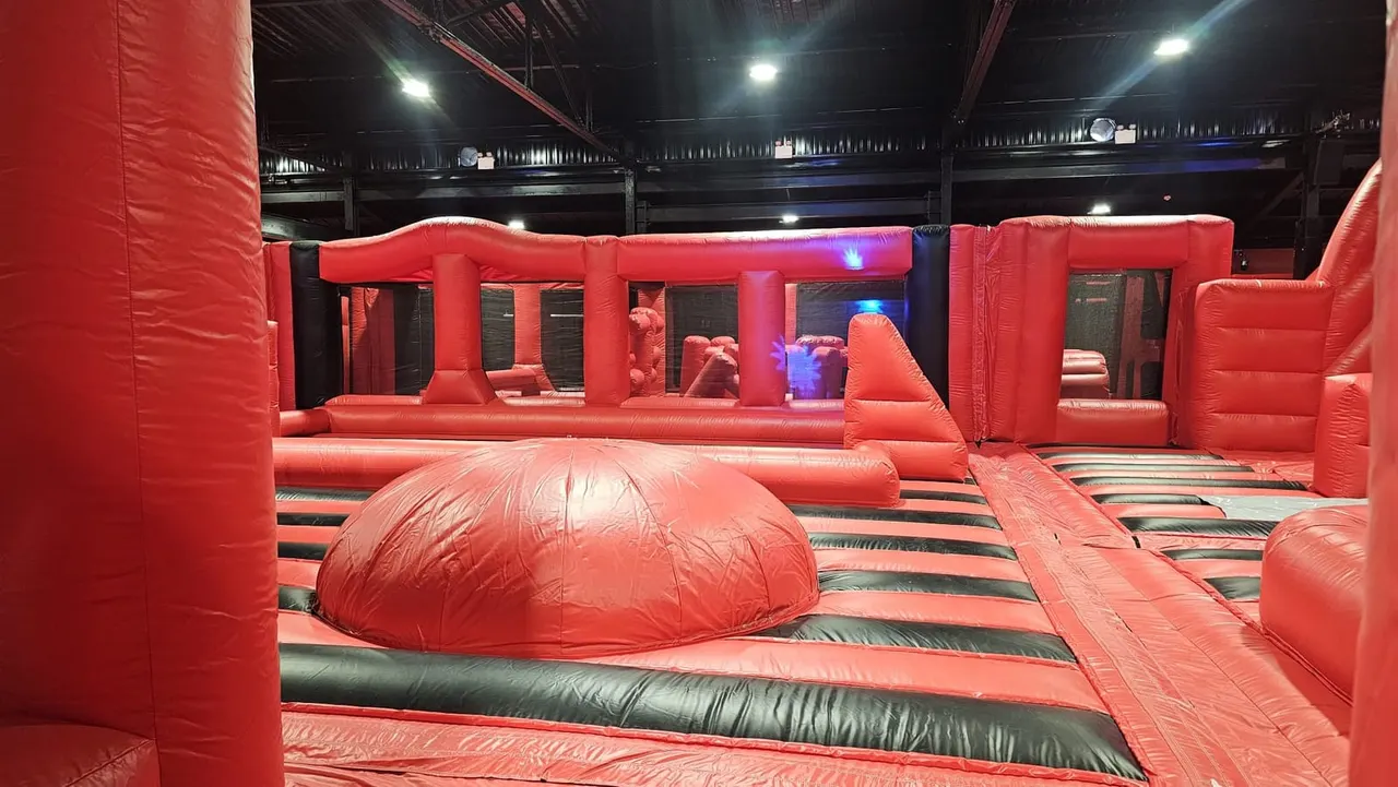 Large red inflatable play arena with obstacle course features, dome structures and LED lighting under industrial ceiling.