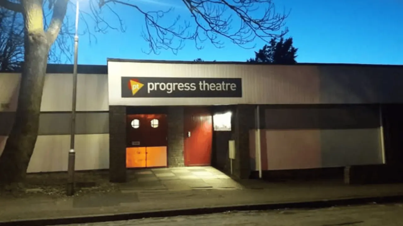 Modern theatre building exterior with illuminated signage and orange entrance doors at dusk.