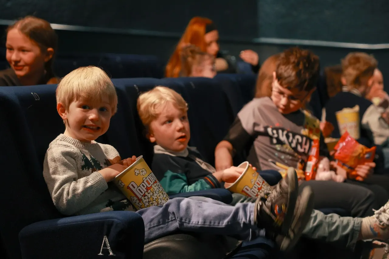 Children sat in cinema seats holding popcorn boxes whilst watching a film.