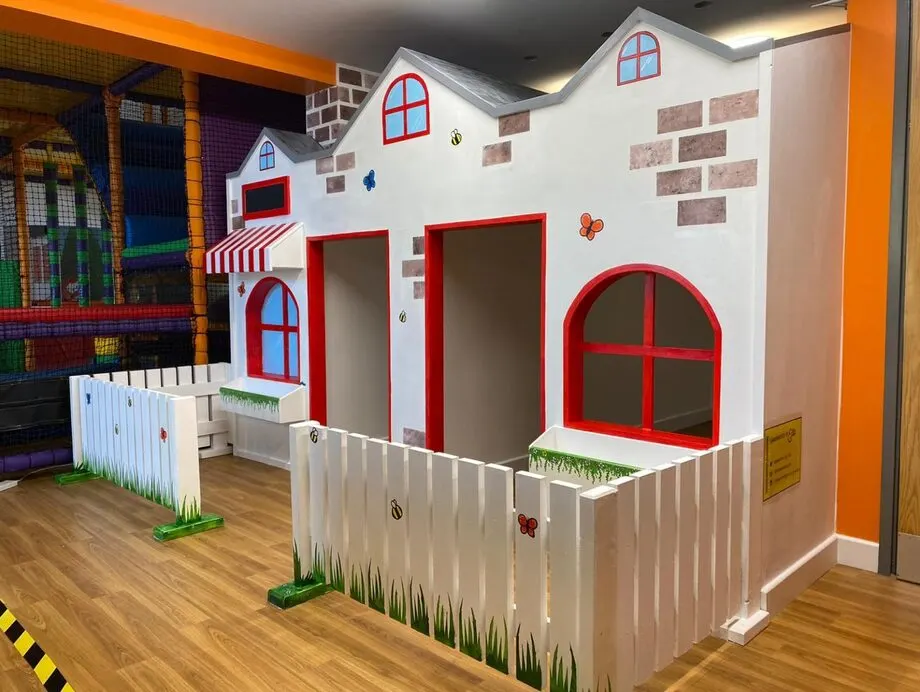 Brightly decorated play house with red doors and windows, white picket fence and soft play equipment in an indoor children's play area.