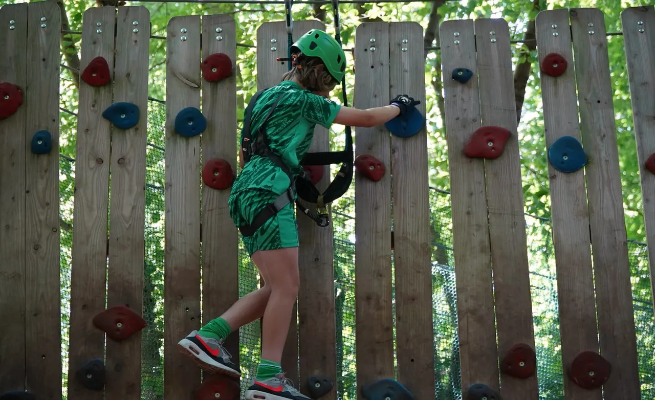 Child in green helmet and harness climbing wooden wall with coloured handholds in a tree canopy course.