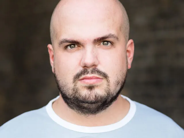 This is a photograph, so I'll provide alt text:

Man with shaved head and dark beard wearing light blue t-shirt, looking directly at camera.