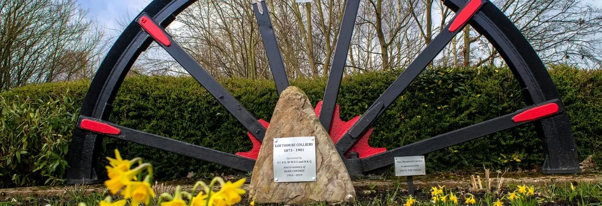 Large black pit wheel with red details and memorial stone set in countryside with yellow flowers and hedges.