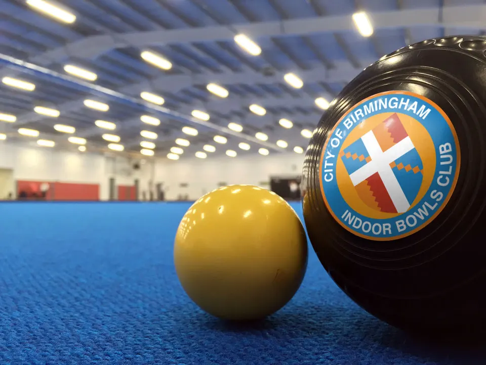 A yellow bowl and black bowls bowl with club emblem on a blue indoor green under bright ceiling lights.