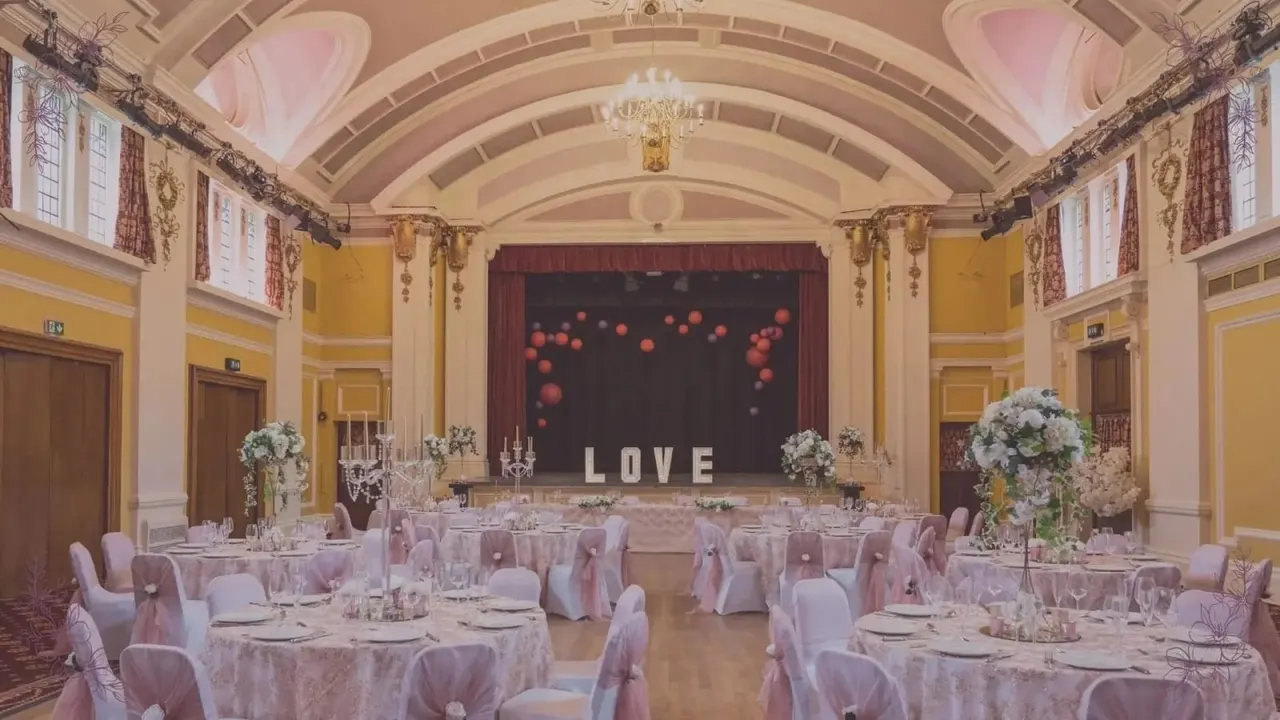 Elegant ballroom decorated for a wedding reception with round tables set with pink linens, floral centrepieces, and place settings facing a stage with 'LOVE' lettering and red balloon decorations.