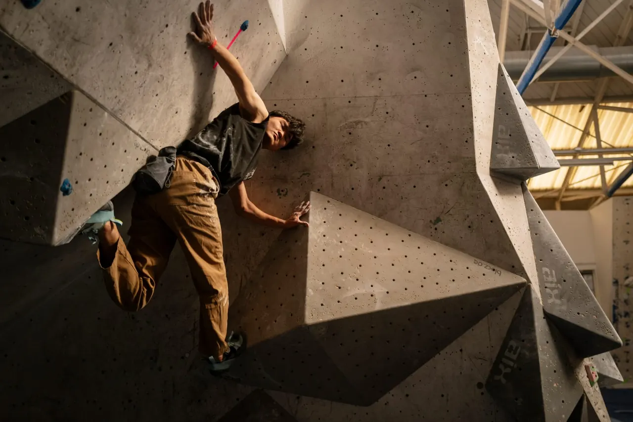 A climber ascending an indoor rock climbing wall with coloured hand holds, wearing a harness and safety equipment.