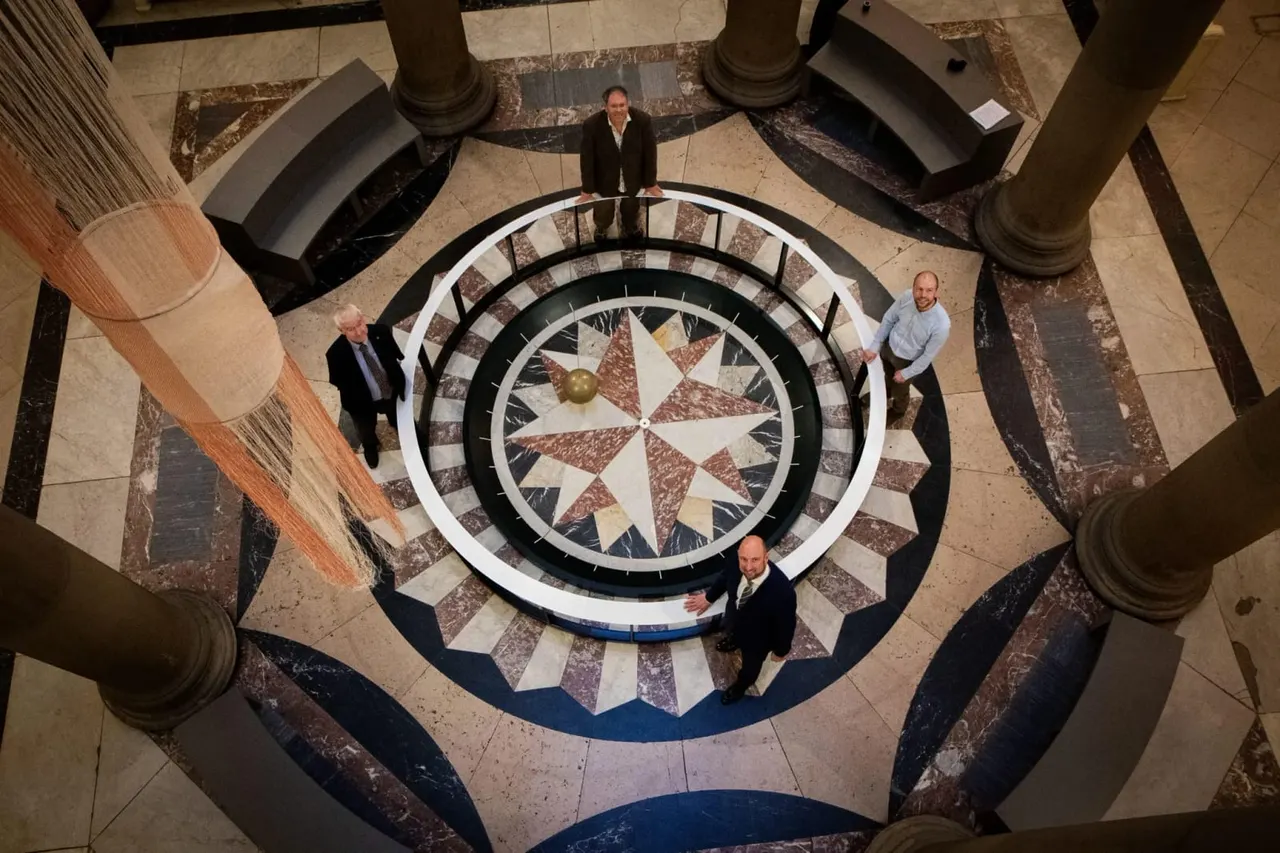 Four people standing around a geometric star pattern floor viewed from above in a grand interior space with columns.