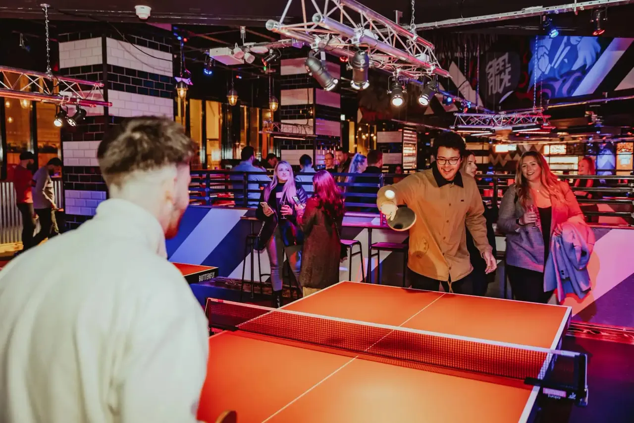 People playing table tennis in a brightly lit indoor entertainment venue with colourful lighting and stage rigging overhead.