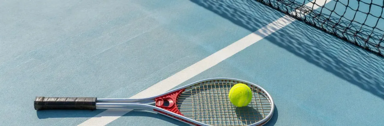 Tennis racket and ball on a blue court with net visible in the background.
