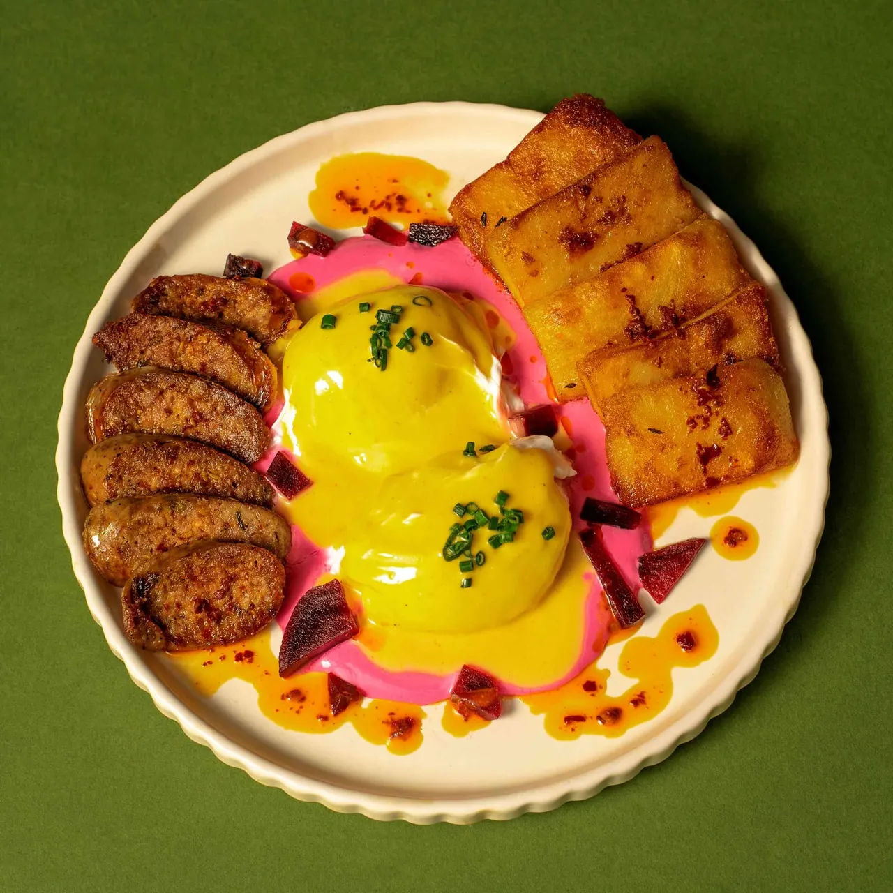 Colourfully plated breakfast dish with fried eggs, toasted bread soldiers, sausages and beetroot on a green background.