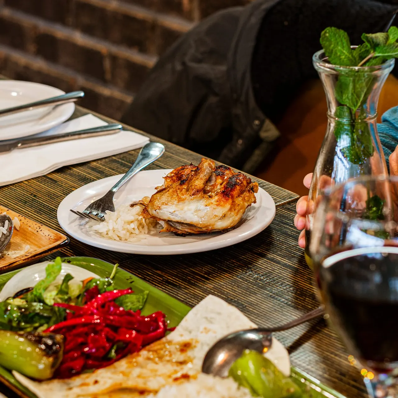 Dining table set with roasted chicken breast and rice, fresh salads with red peppers, and mint water, at an indoor restaurant.