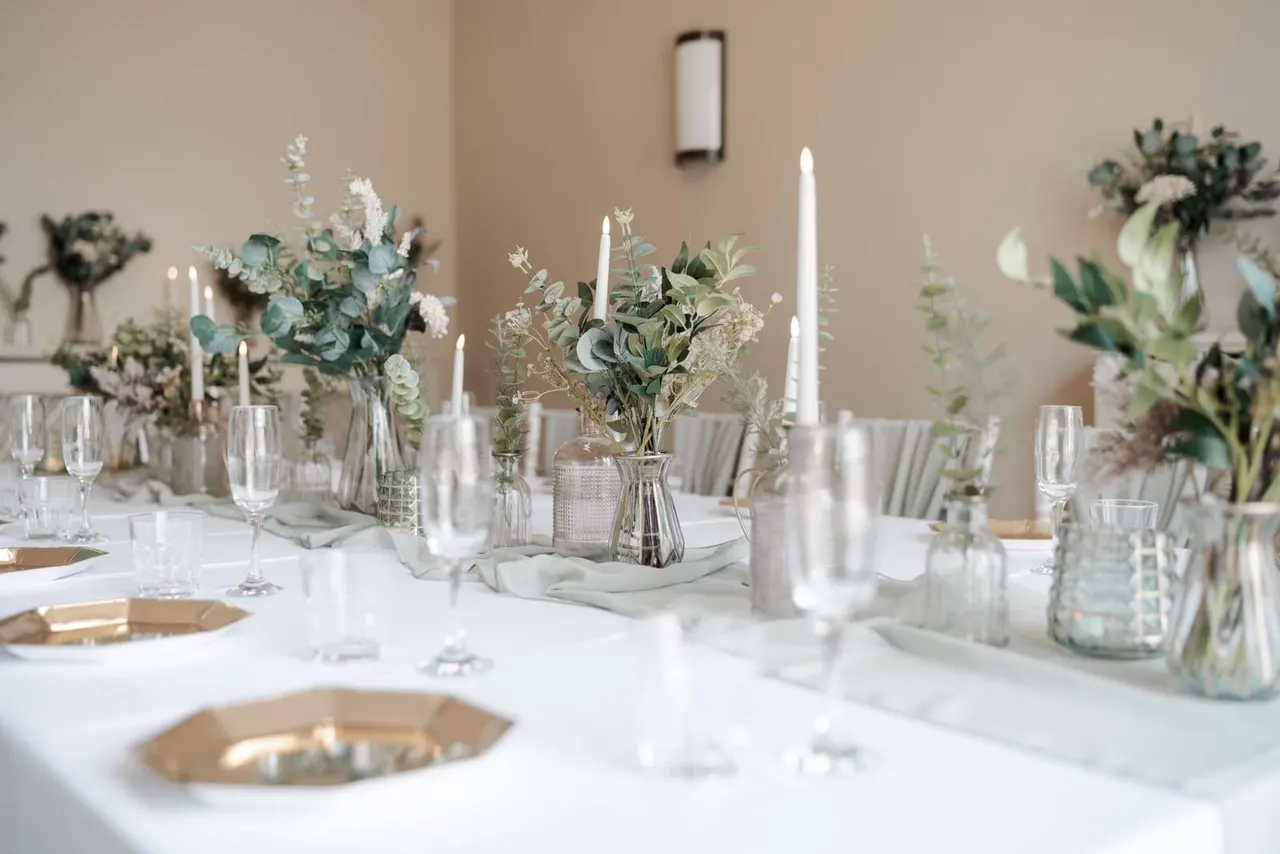 Elegantly dressed dining table with white linens, greenery arrangements, candles, and place settings in a neutral interior space.