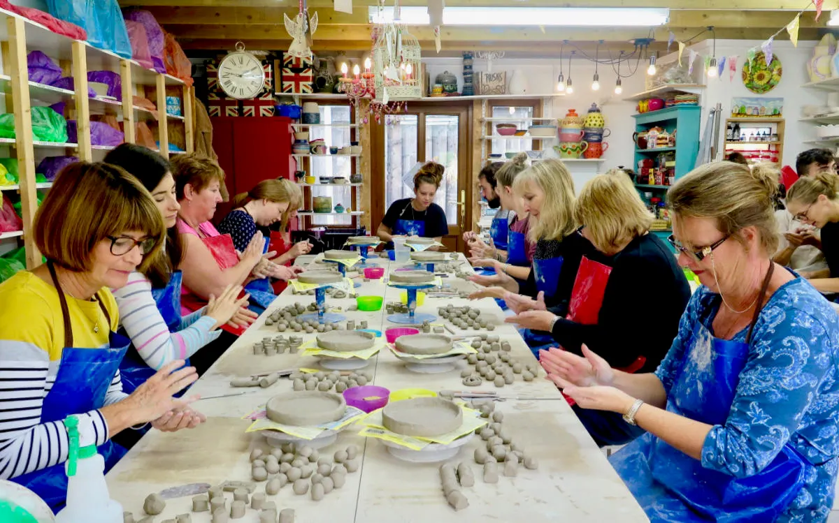 Adults and children work at pottery wheels and hand-mould clay at tables in a bright craft studio.