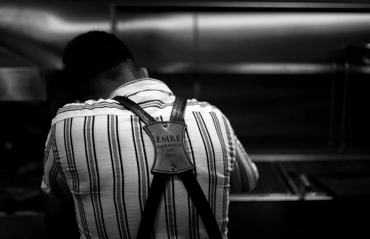 Person wearing a striped shirt and branded apron viewed from behind in a restaurant kitchen.