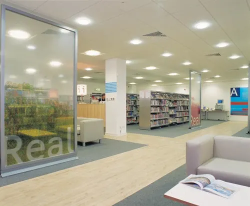Modern library interior with seating area in foreground and open-plan shelving visible through glass partition.
