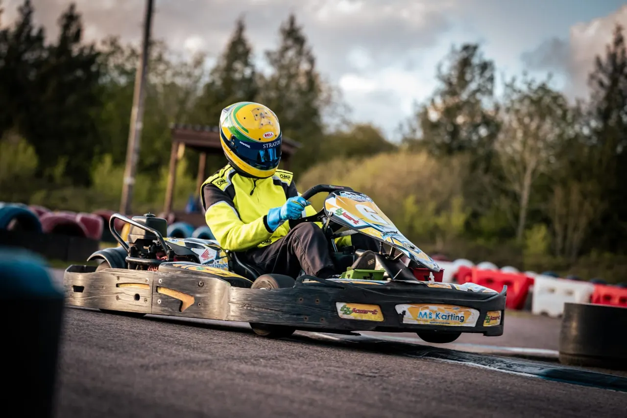 Racer in yellow helmet and suit driving a go-kart on an outdoor track lined with coloured bumpers and conifers.