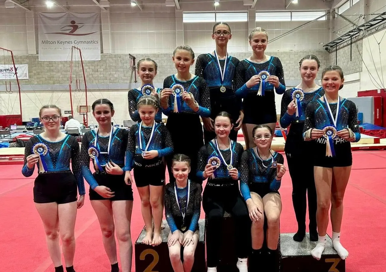 Young gymnasts in sparkly leotards displaying medals at a competition in a gymnasium with red mats.