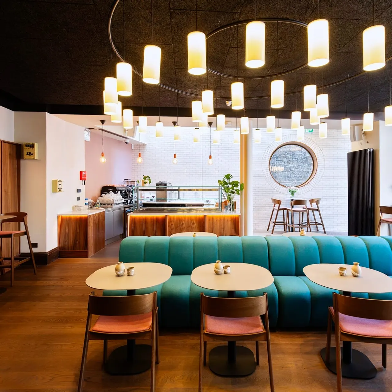 Modern café interior with green curved seating booths, round tables, pendant lighting, and wood-panelled counter service area.