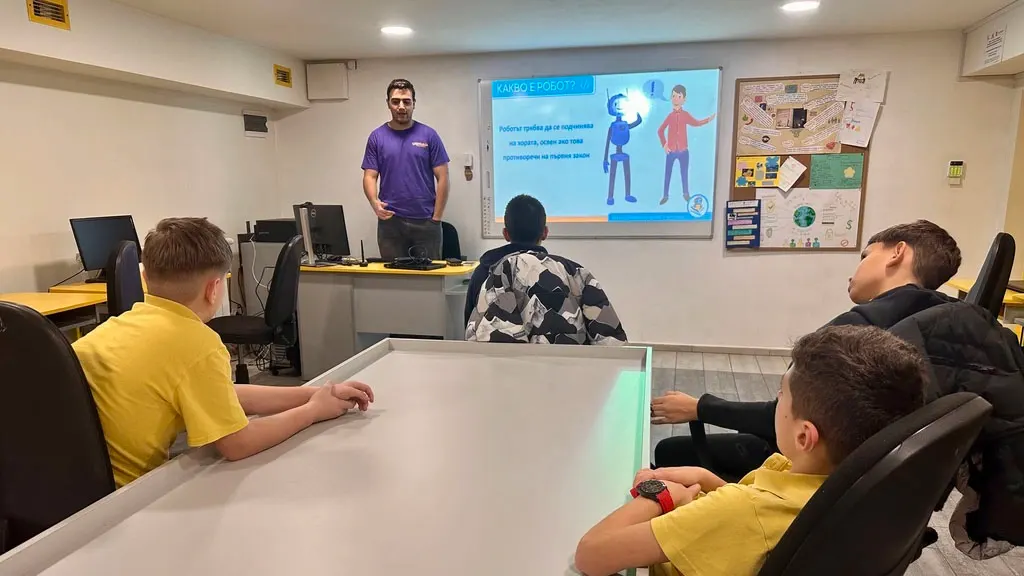 An instructor in purple stands by a projector screen whilst several young people in yellow shirts sit at a table during a coding class.