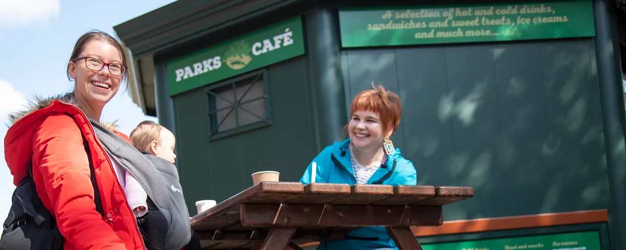 Woman with baby and young child smiling outside a parks café at a picnic table.