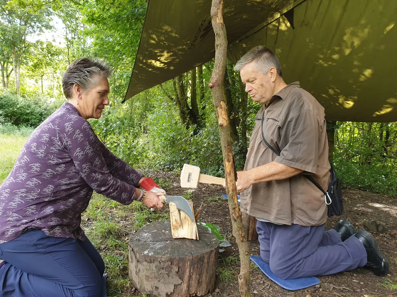 Two adults practising wood carving with chisels and mallets under a canvas shelter in a woodland setting.