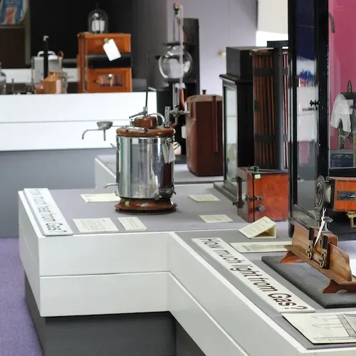 Museum display of vintage gas appliances and heating equipment including a pressure vessel and wooden-cased devices on white display cases.