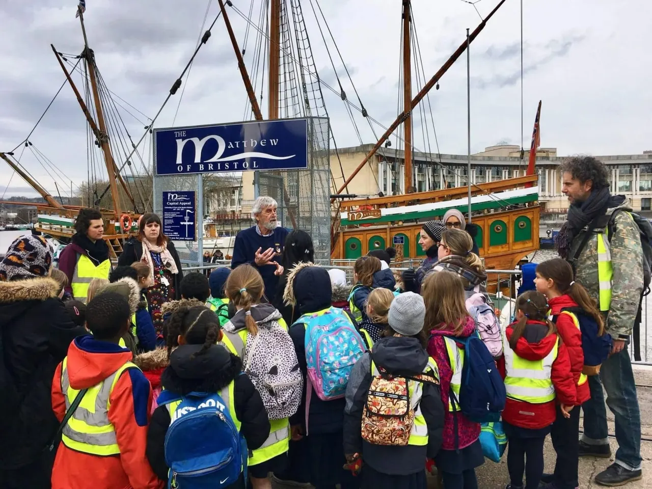 School group wearing high-visibility vests listens to guide beside the wooden sailing ship Matthew of Bristol.