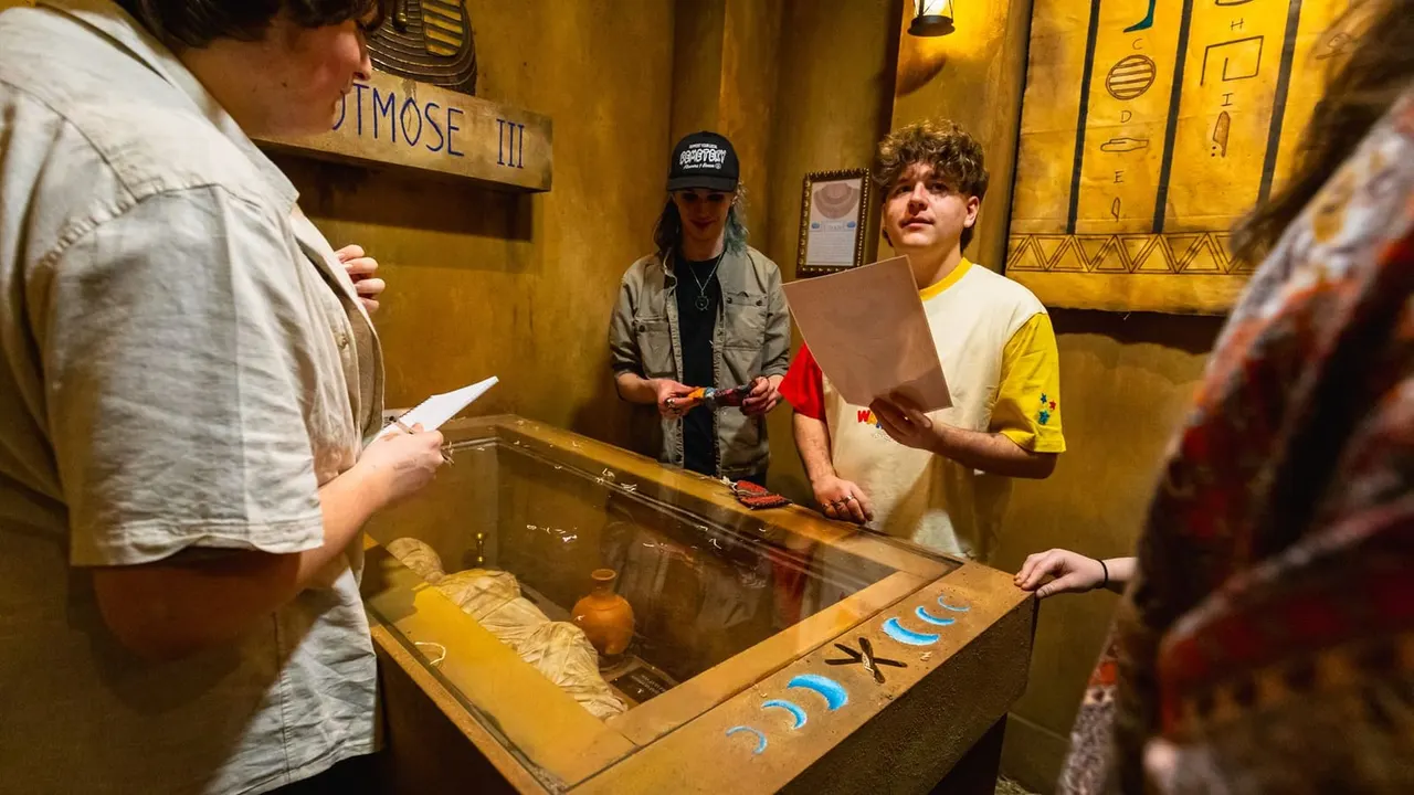 Young people gathered around a display case in an Egyptian-themed room with hieroglyphic decorations and a mummy exhibit.