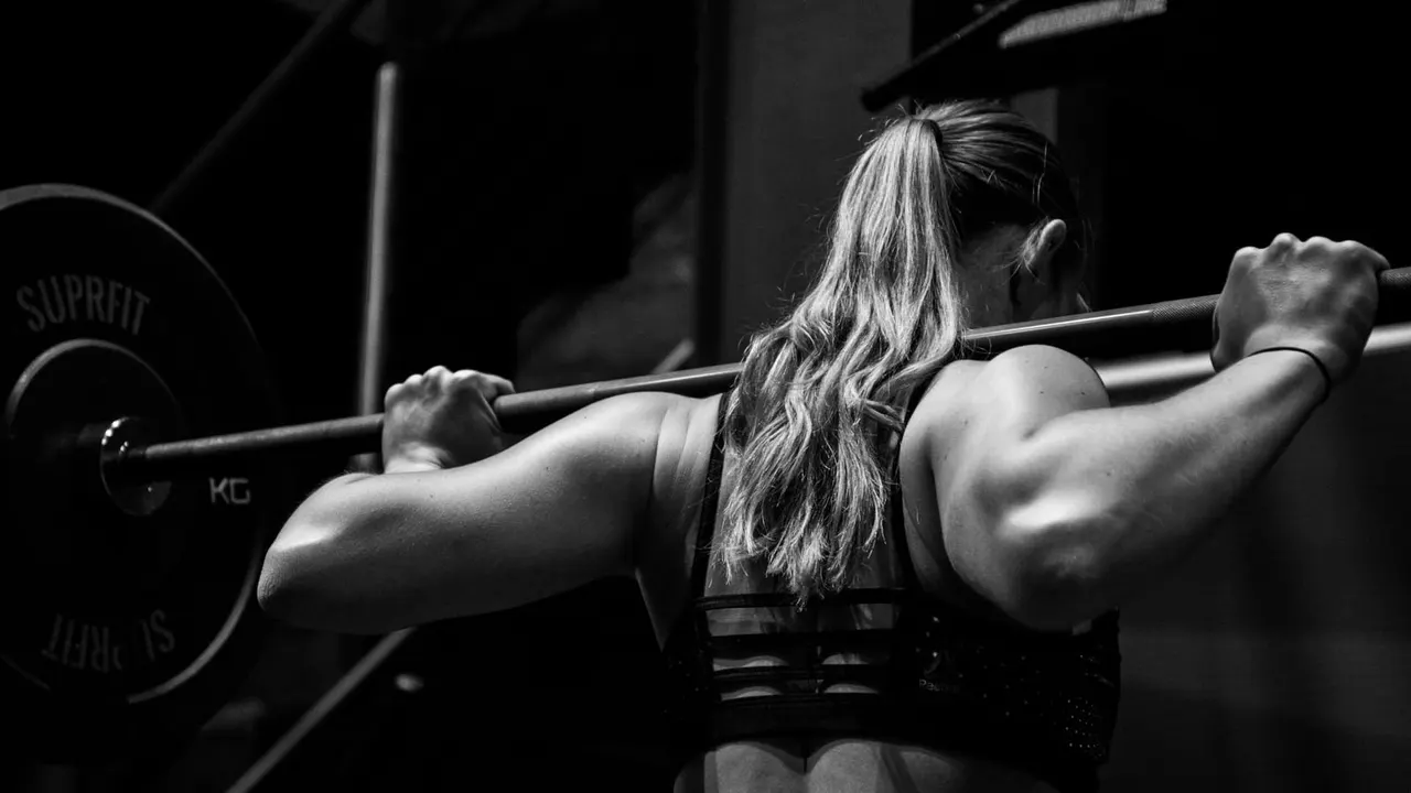 Woman performing a barbell squat exercise in a gym, viewed from behind in black and white.