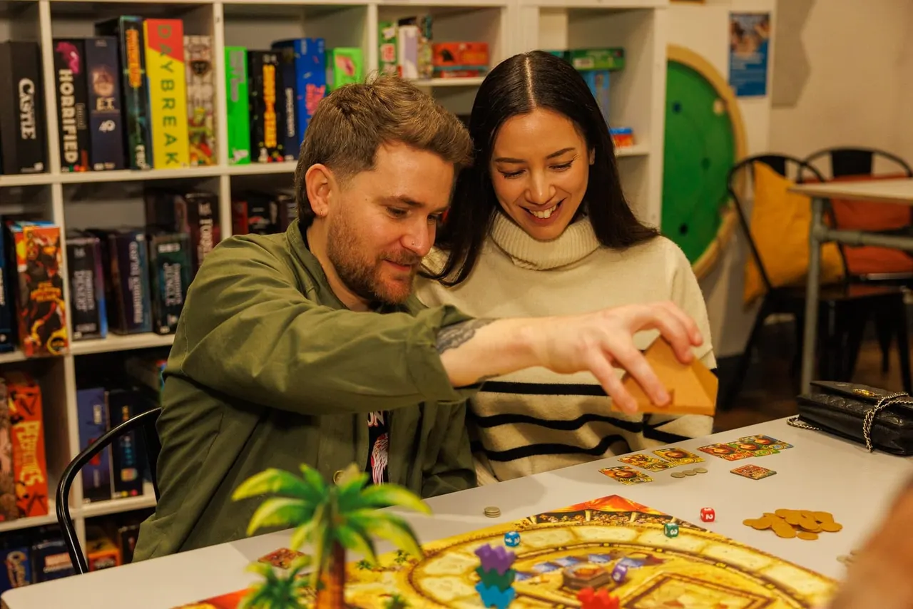 Two people playing a board game together at a table in a games room with shelves of boxes behind them.