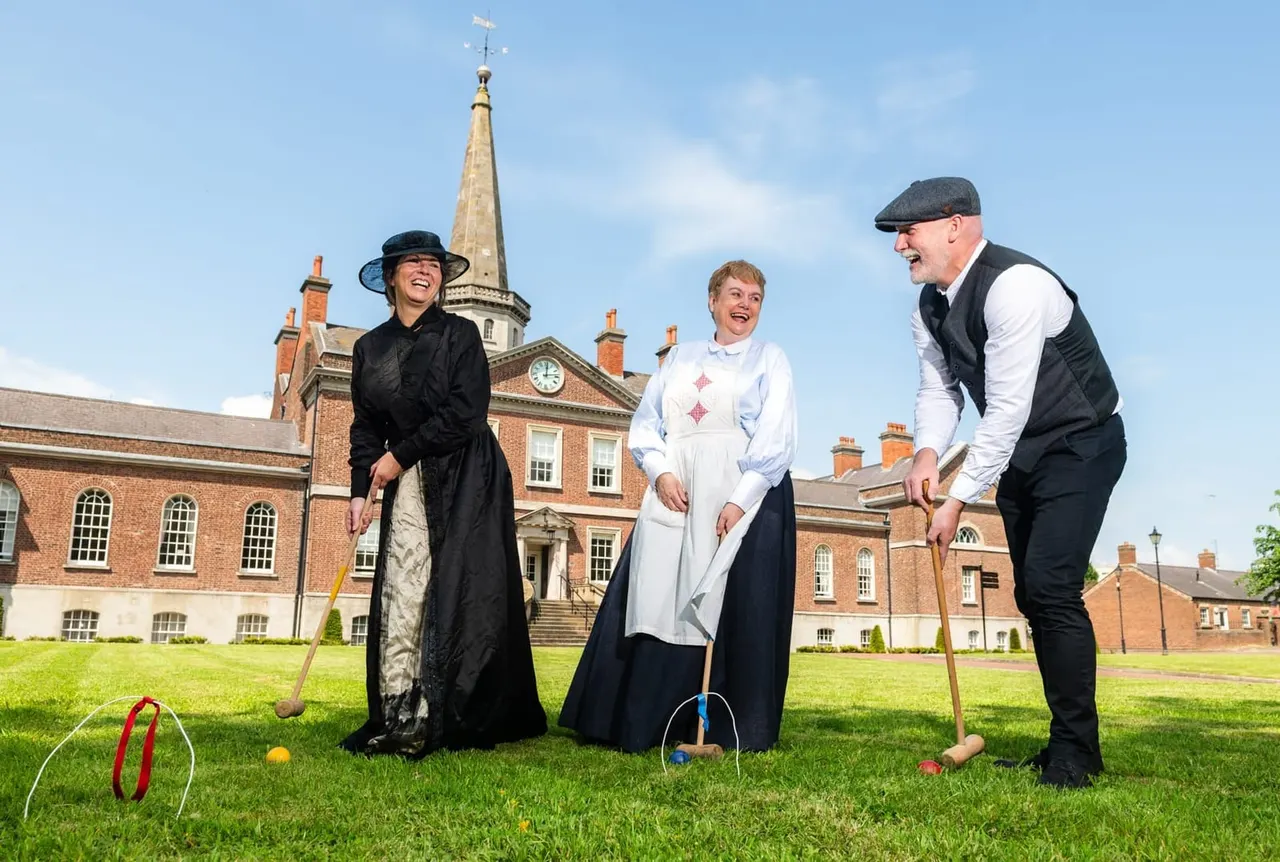 Three people in period costume playing croquet on a lawn in front of a Georgian brick building with a church spire.