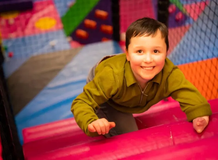 Child smiling while playing on a colourful soft play structure with foam padding and climbing elements.