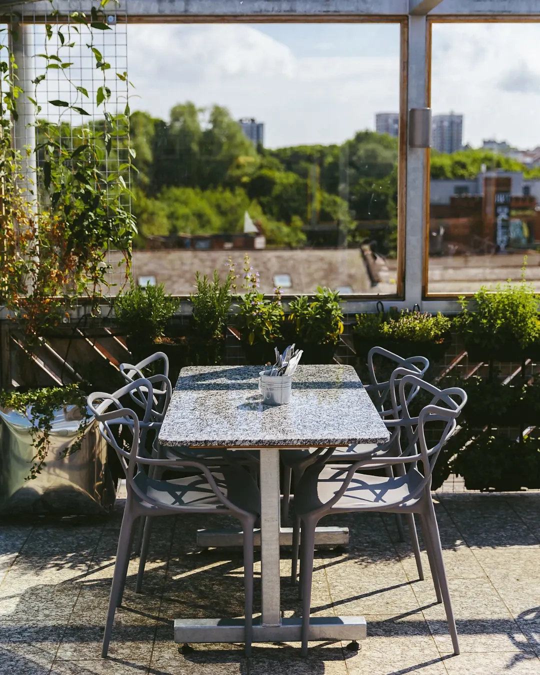 Outdoor seating area with a table and chairs on a rooftop terrace overlooking a river, green trees and urban skyline.