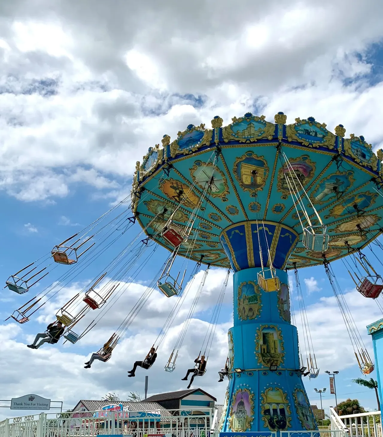 Ornate blue and gold carousel swing ride in motion with riders soaring outwards against a cloudy sky.