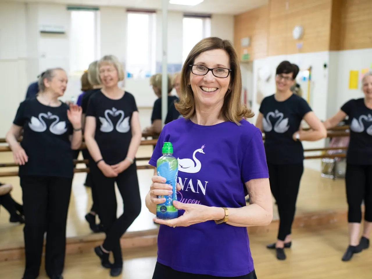 Woman in purple t-shirt holding water bottle smiles at camera whilst dancers in black t-shirts with swan motifs stand behind her in a studio.