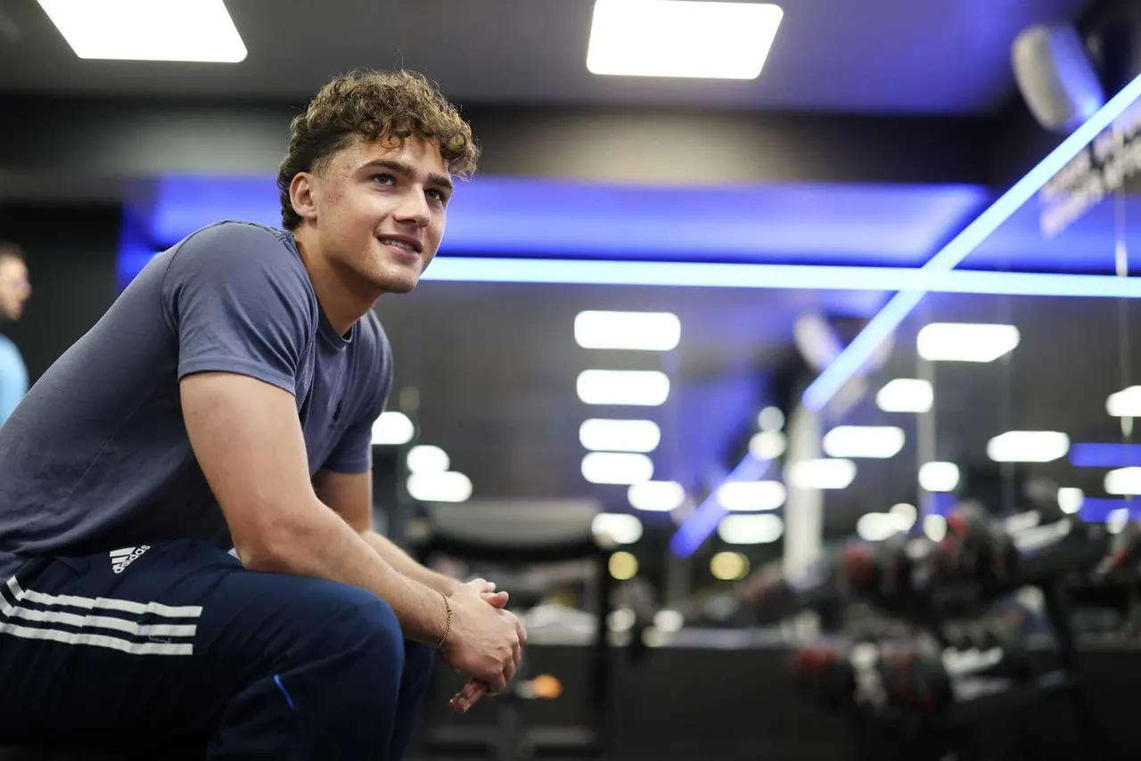 Young man crouching inside a modern sports facility with blue lighting and other people in the background.