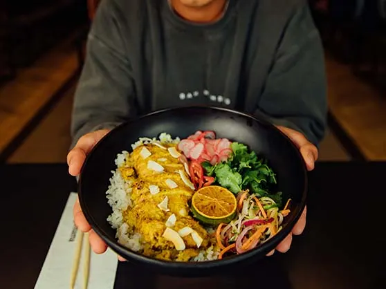 Person holding a bowl of Asian noodle dish with egg, vegetables and garnishes.