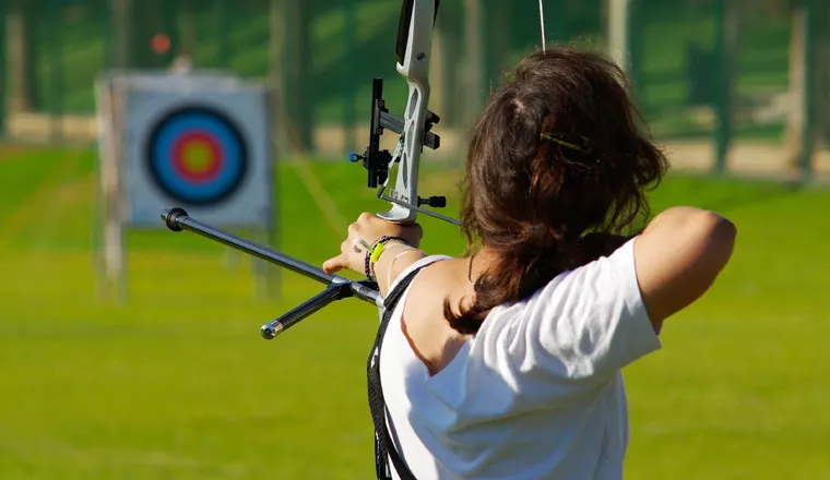 Person drawing a compound bow aimed at a distant target during an outdoor archery session.