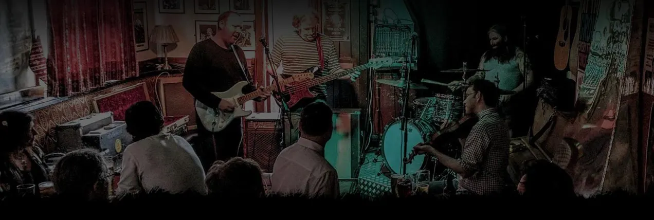 Band performing live in intimate pub setting with audience watching musicians play guitar and drums.
