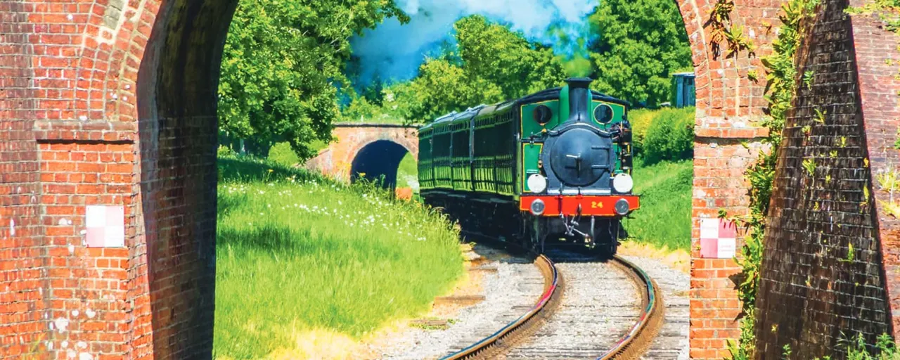 Small heritage railway engine with green and red livery approaching through brick railway arches on a sunny day.