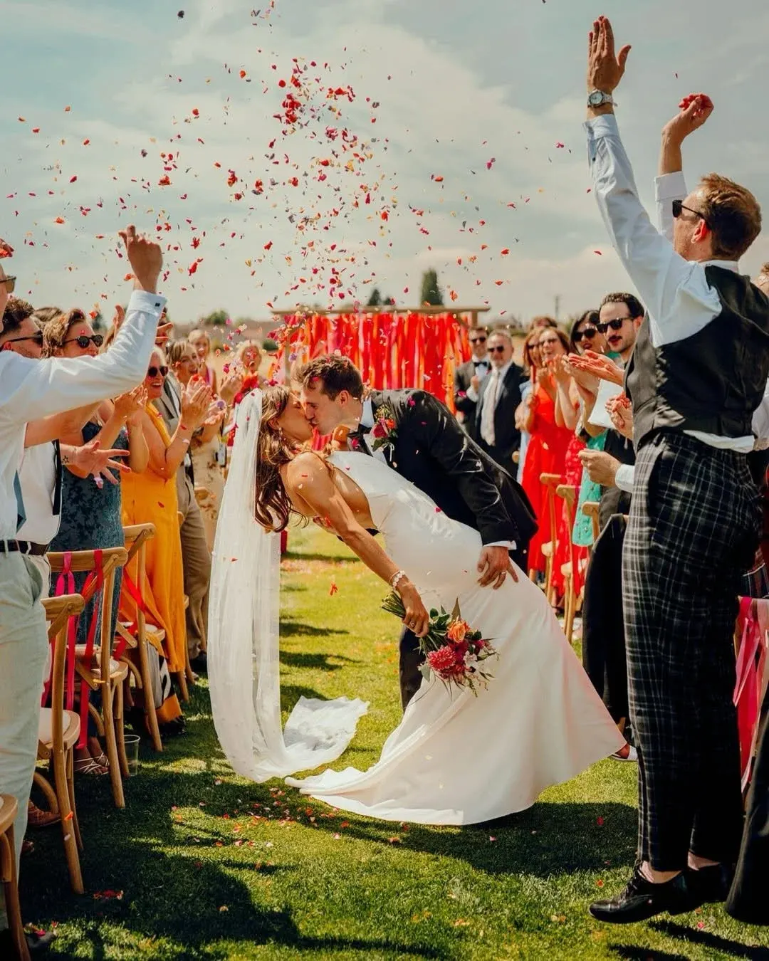 Bride and groom kiss while guests throw red confetti during outdoor wedding ceremony on grass.